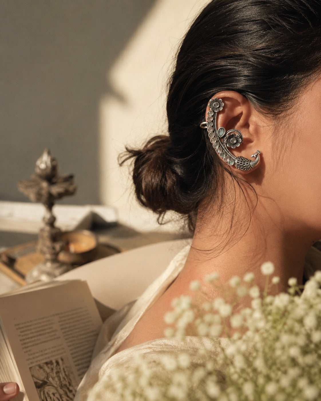 A close-up lifestyle shot of a woman wearing a large, ornate silver crescent-shaped ear cuff with intricate floral detailing. She is shown in profile with her hair in a bun, wearing a cream-colored top, with a soft-focus background featuring a candle, an open book, and white baby's breath flowers.