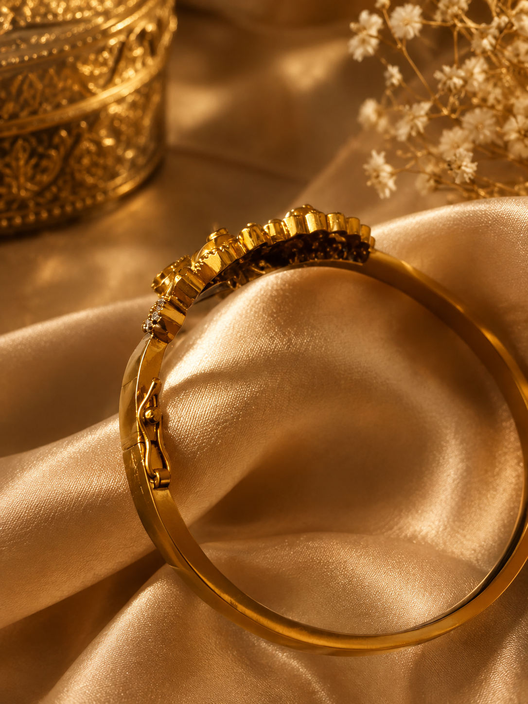 A gold-toned bangle bracelet with a decorative floral crown-like setting, displayed on draped champagne satin fabric with dried flowers and a gold ornate container in the background.