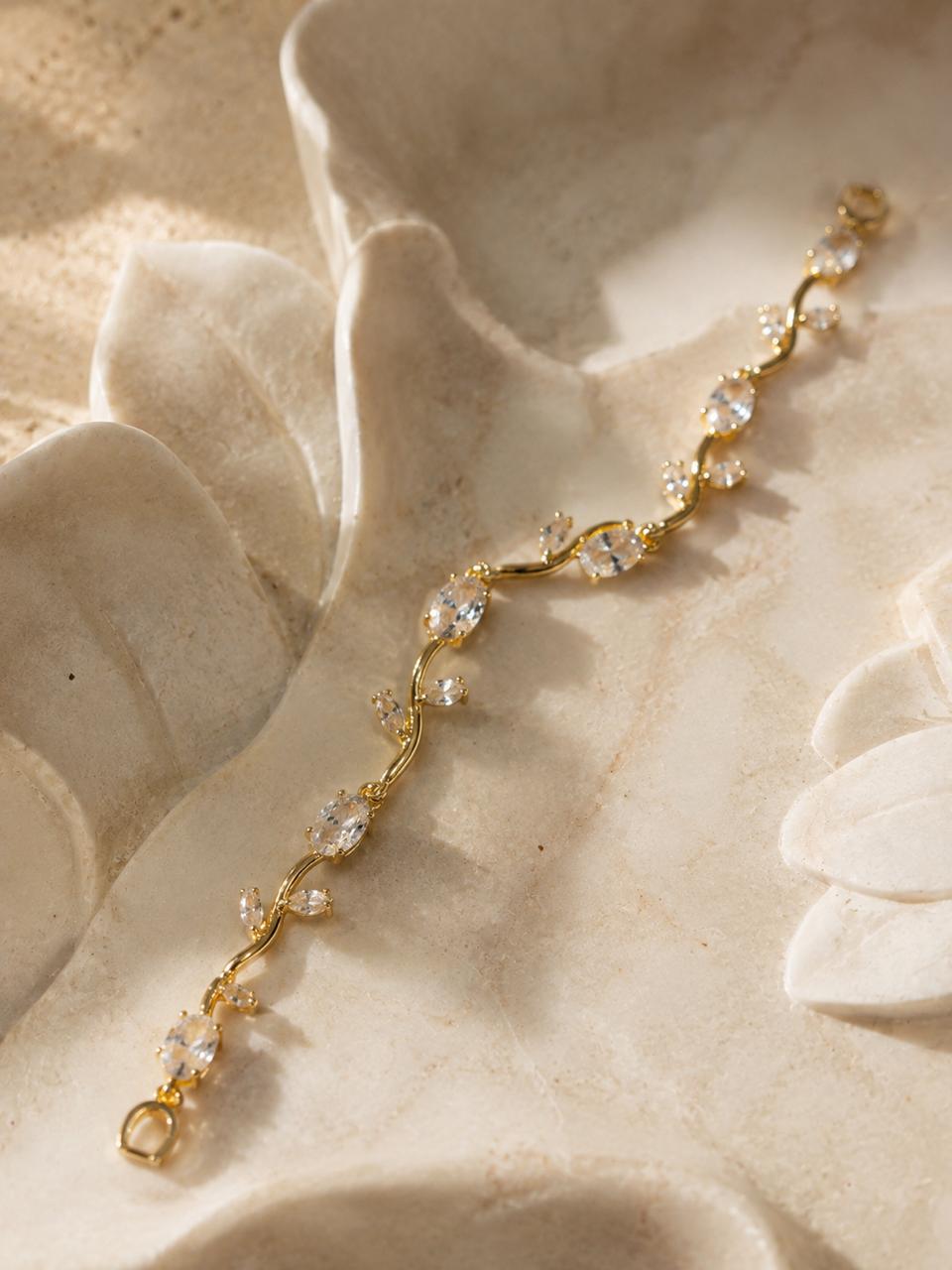A delicate gold vine bracelet adorned with clear, faceted gemstones, displayed on a textured beige stone surface.