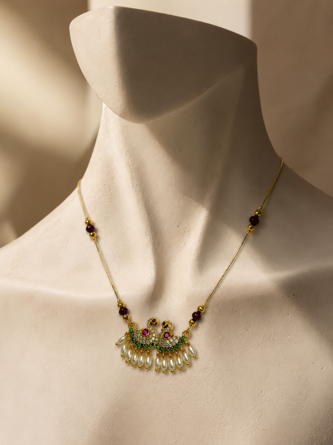 A gold-toned necklace with a decorative pendant displayed on a cream-colored mannequin bust. The necklace features a gold chain, small dark purple beads, green and red stone accents, and a fan-shaped pearl-like pendant at the center.