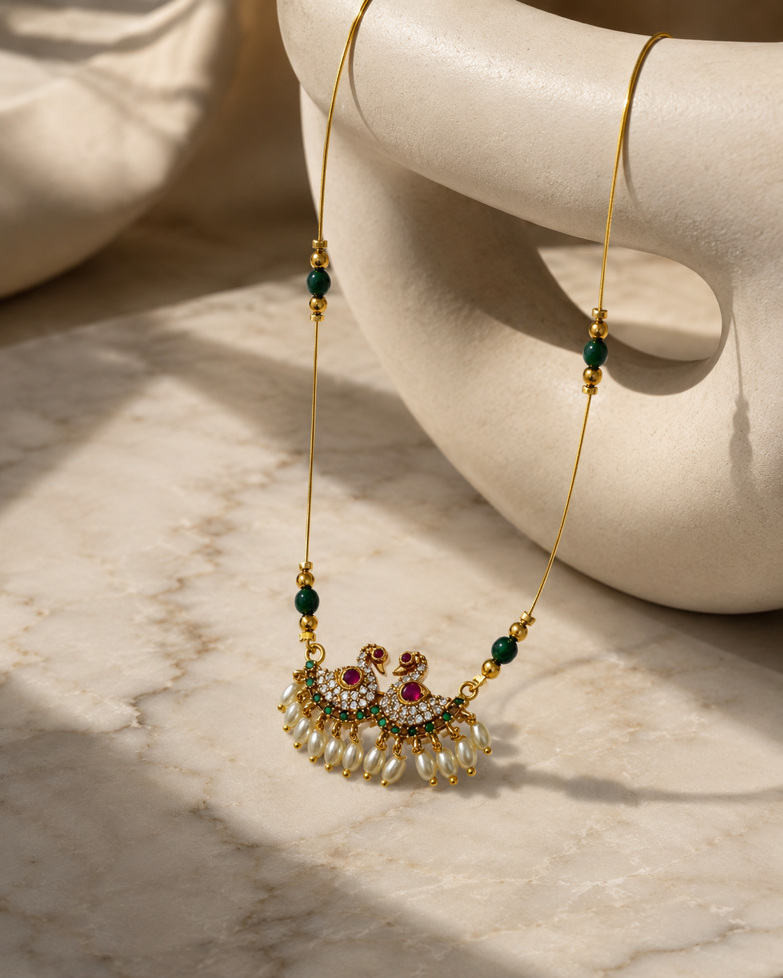 A gold-tone necklace with a decorative pendant featuring white pearls, green beads, and red and white stones, displayed on a cream ceramic stand over a marble surface.