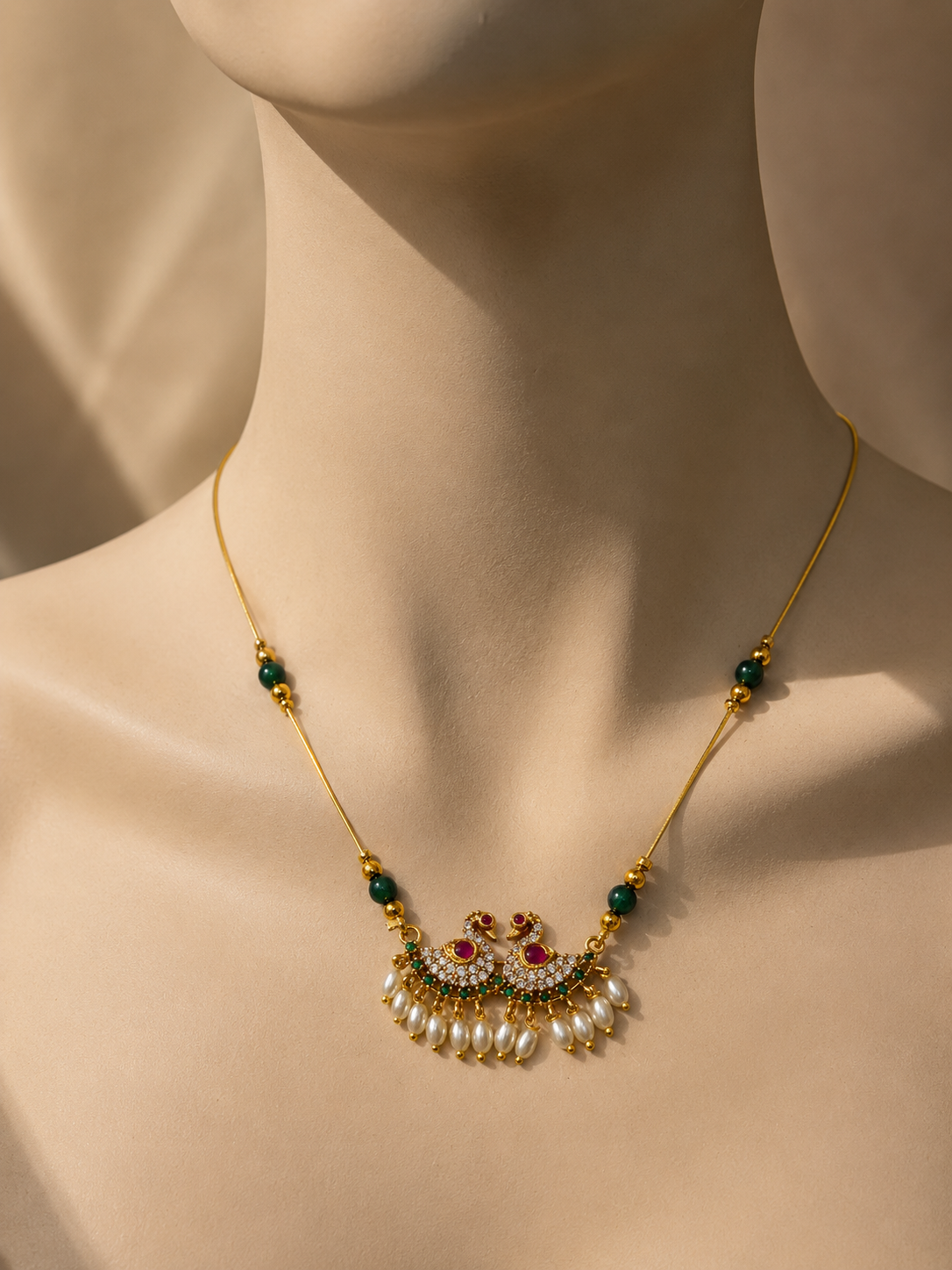 A gold-plated traditional Indian choker necklace with a detailed peacock pendant, set on a beige mannequin bust. The necklace features green stone accents, white pearls, and small red and white stones in the peacock's body.