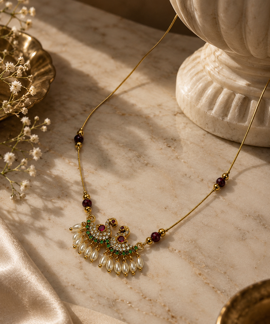 Gold-toned necklace with a decorative pendant featuring white pearl-like drops, green stones, and dark red beads, styled on a beige marble surface with soft decorative props.