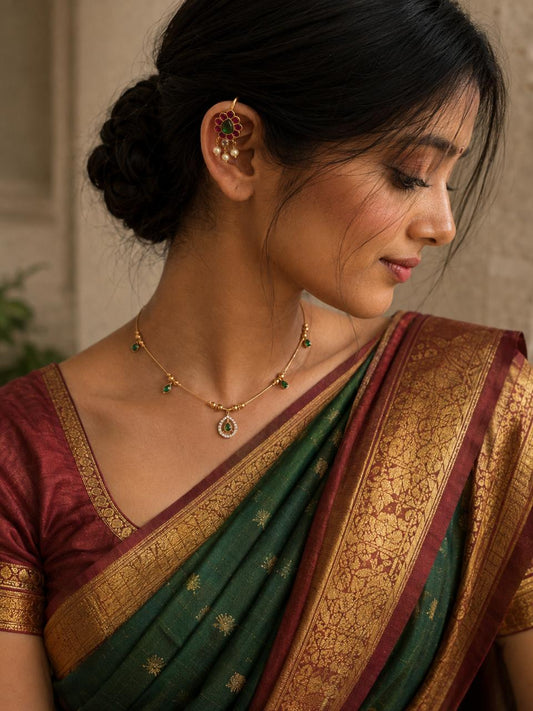 A woman wearing a green and maroon silk saree with a gold zari border, paired with a gold necklace and a gold ear cuff with red stones, shown in a close-up profile view.