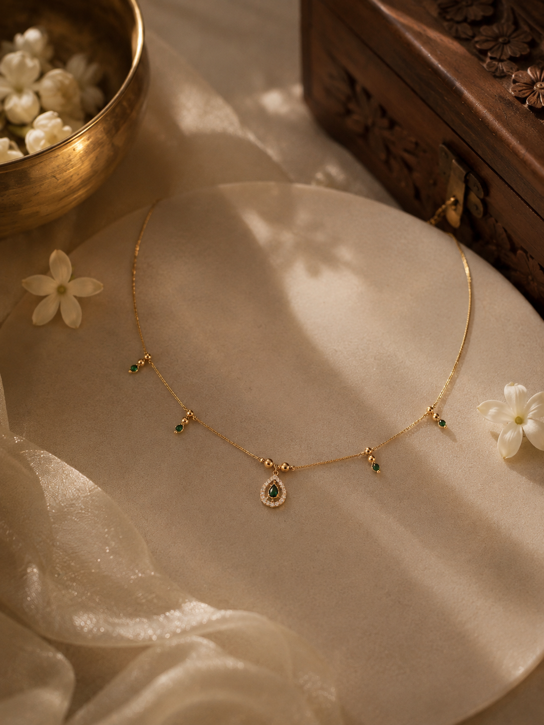 A delicate gold chain necklace with a teardrop-shaped pendant featuring green stones, styled on a beige fabric surface with white flowers and a brass bowl in the background.
