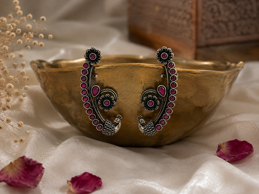 A pair of ornate silver-toned earrings with pink gemstone accents and peacock motifs, displayed on a gold-toned bowl over white fabric with scattered rose petals.