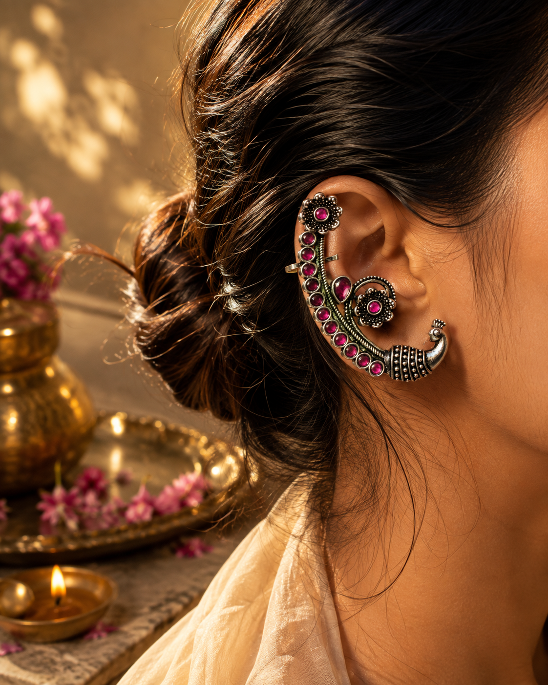 A close-up lifestyle shot of a woman's ear adorned with an intricate silver-toned traditional Indian ear cuff decorated with pink gemstones, set against a warm, softly lit background with traditional decor.
