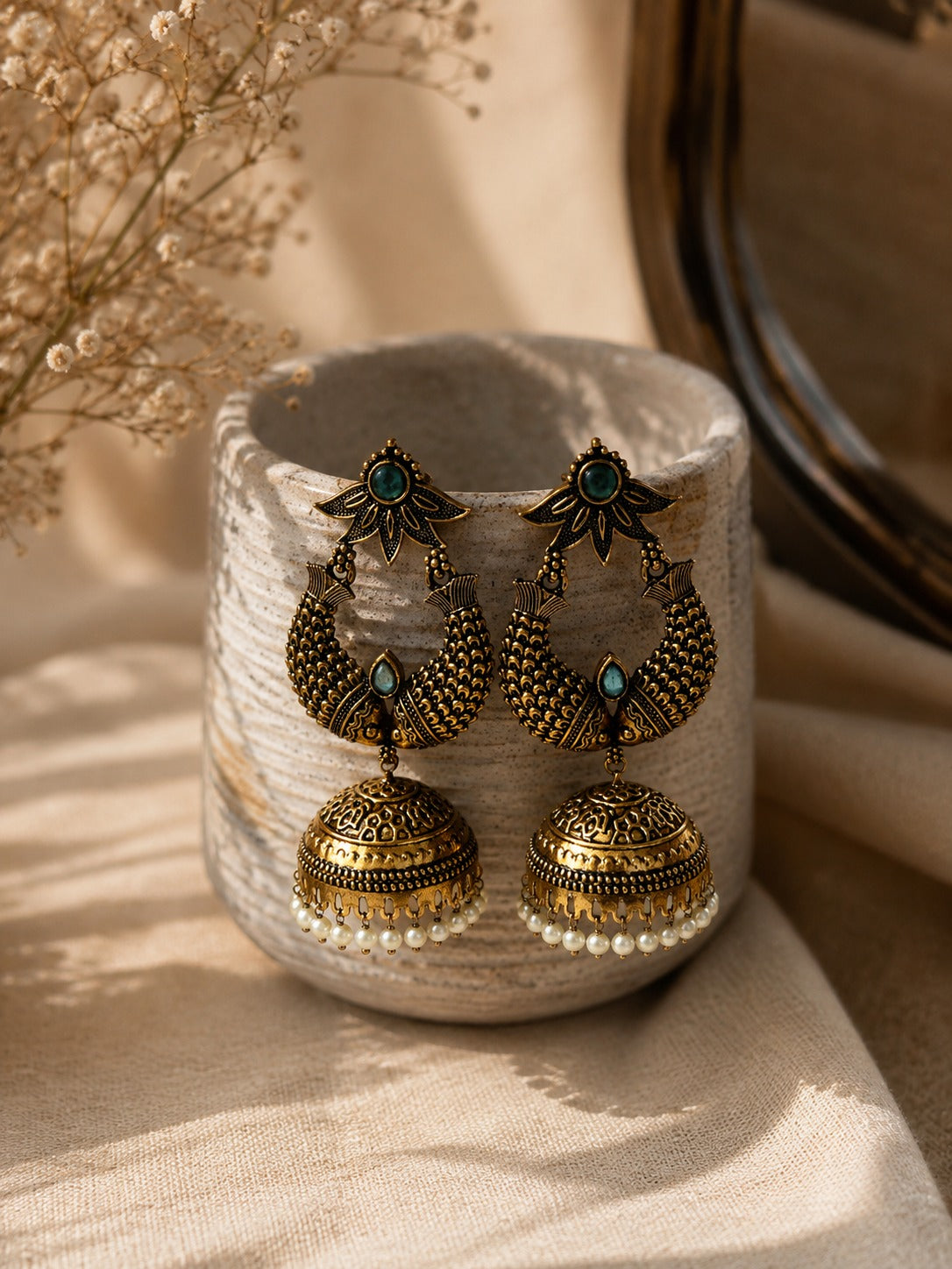 A pair of ornate gold-toned jhumka earrings with turquoise blue stones and small white pearl bead drops, displayed on a textured ceramic cup with neutral fabric and dried flowers in the background.