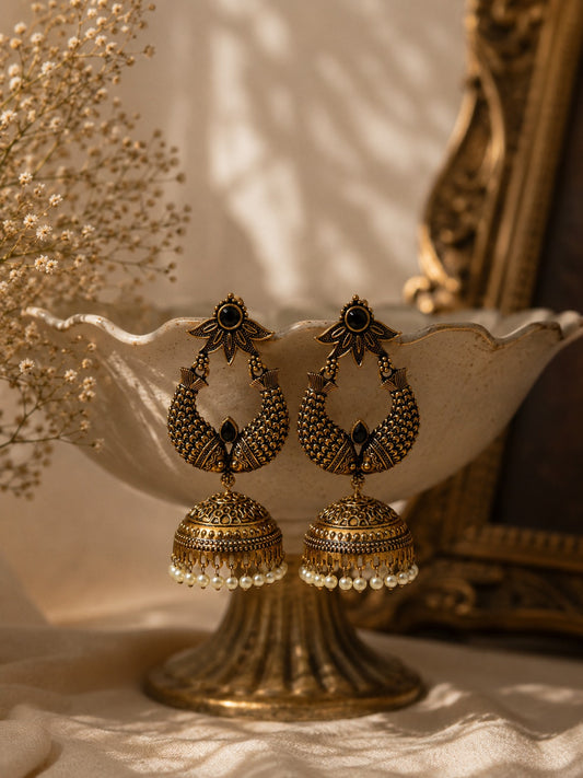 A pair of ornate gold-toned Indian jhumka earrings with black enamel details and small pearl bead drops, displayed on a vintage cream-colored pedestal bowl with warm, shadowed lighting.