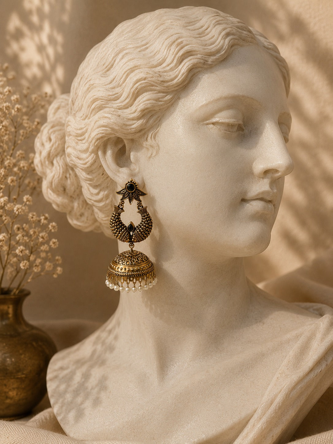 A close-up of a classical marble bust wearing a dangling ornate earring. The earring has an antique gold-toned filigree design with a dark top stone and a beaded drop.