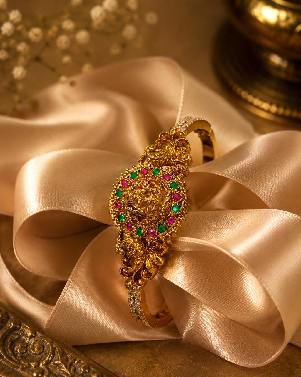 An ornate gold ring with a detailed filigree design, accented with green and pink gemstones, displayed on a beige satin ribbon.