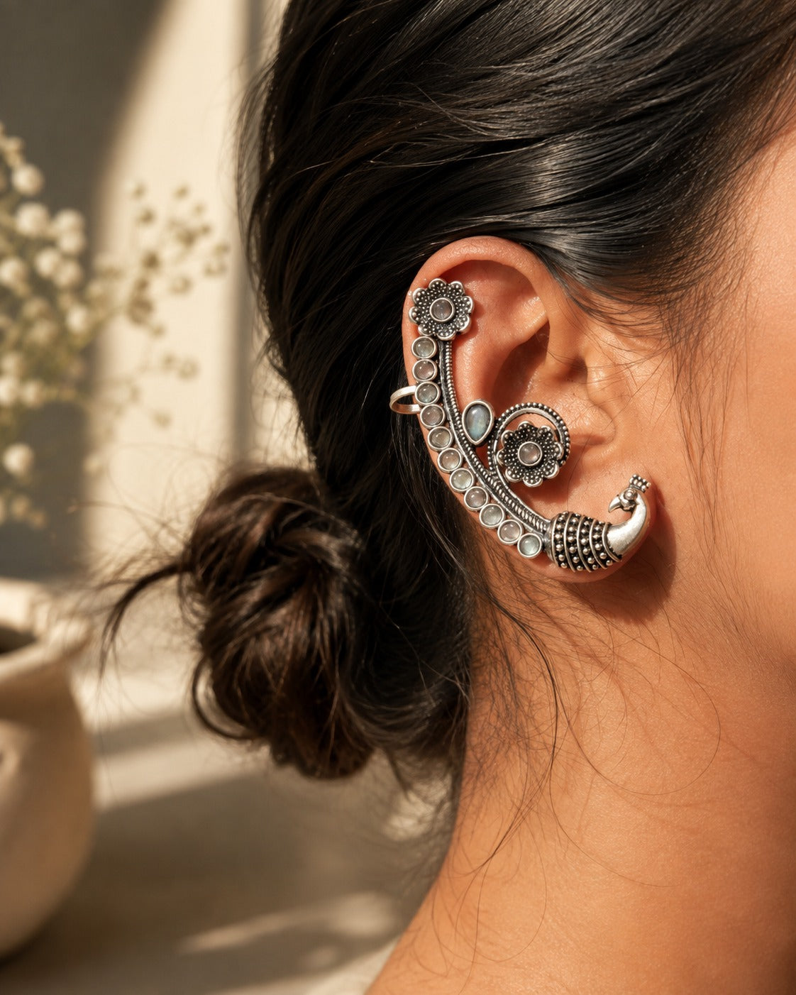 A close-up of a person's ear wearing an ornate silver crescent-shaped ear cuff with floral and geometric details, set against a softly blurred warm-toned indoor background.
