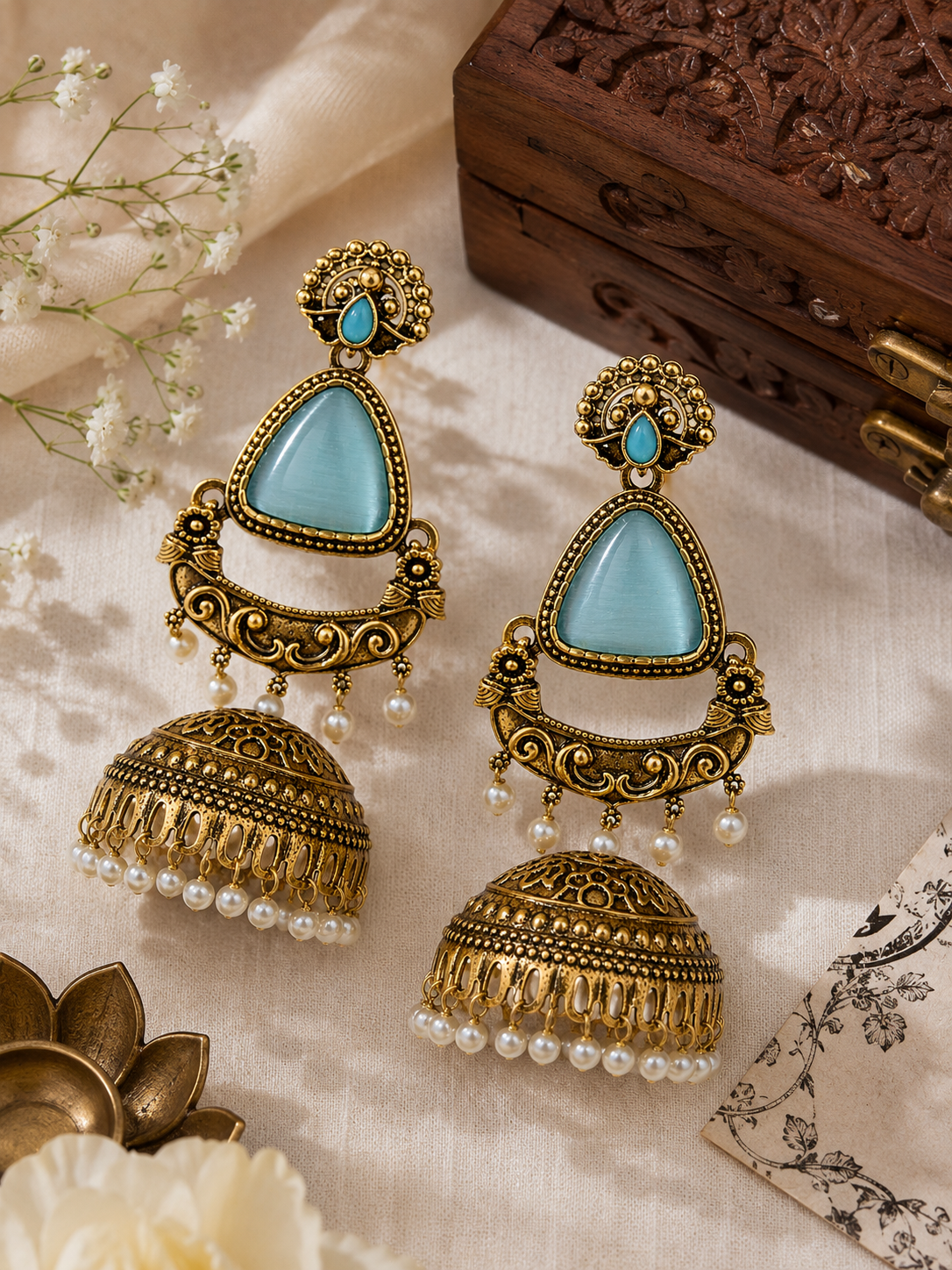 A pair of ornate gold-toned jhumka earrings with light blue stone accents and dangling pearl beads, arranged on a cream fabric surface with decorative floral and wooden props.