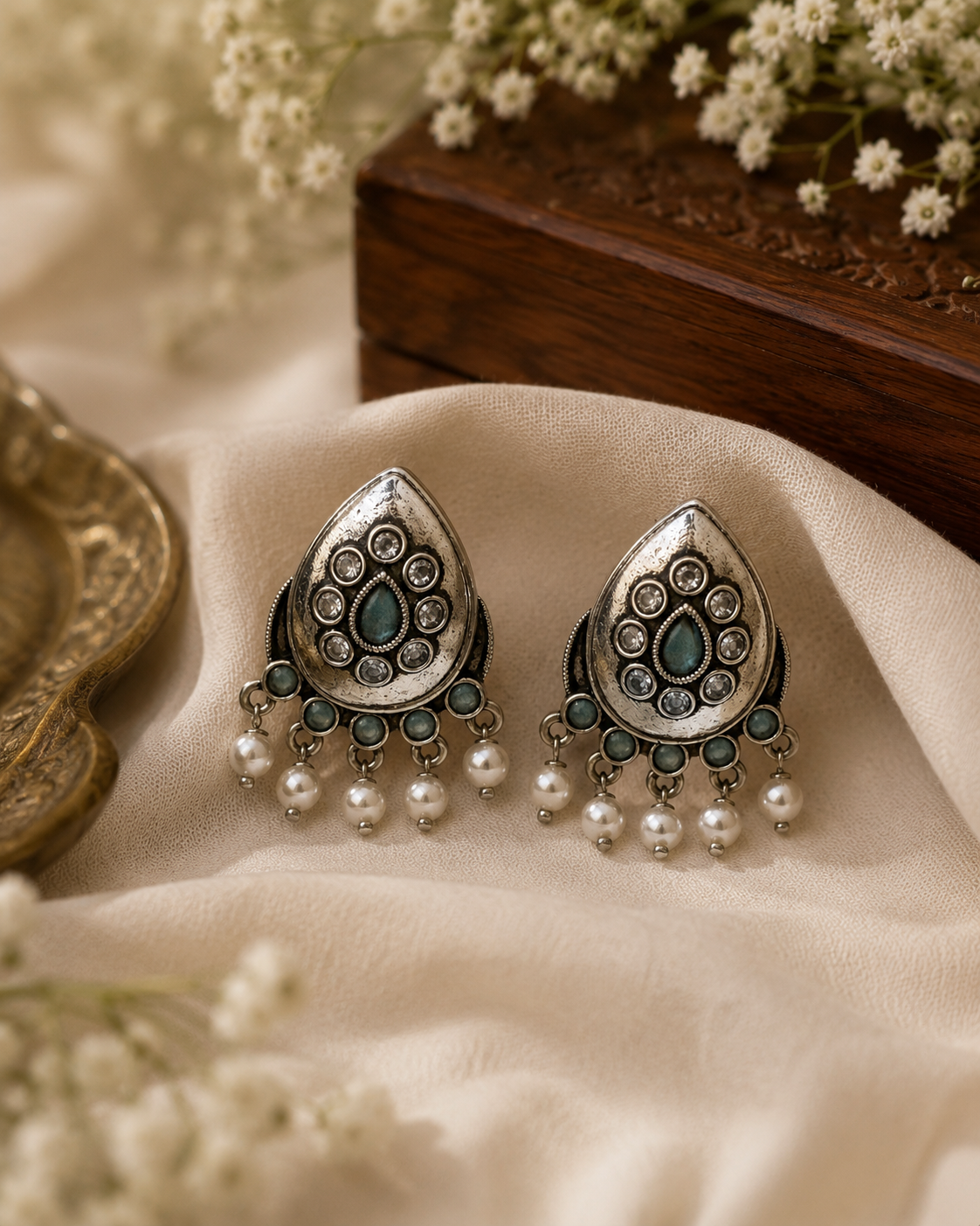 A pair of ornate silver-toned Indian-style earrings with teal blue stones and dangling white pearls, displayed on draped cream fabric with decorative flowers and a wooden box in the background.