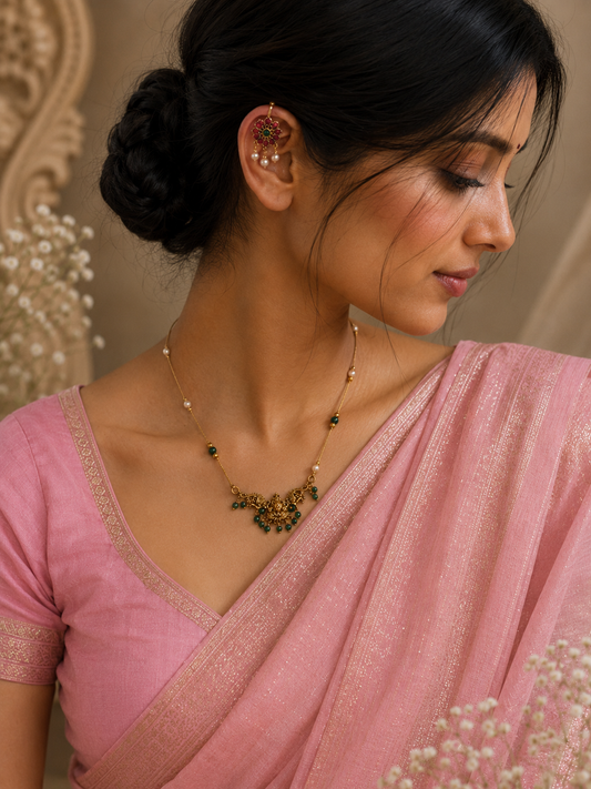 A woman wearing a pink saree and blouse with a gold-toned necklace and matching ear cuff, shown in a close-up portrait against a soft beige background.