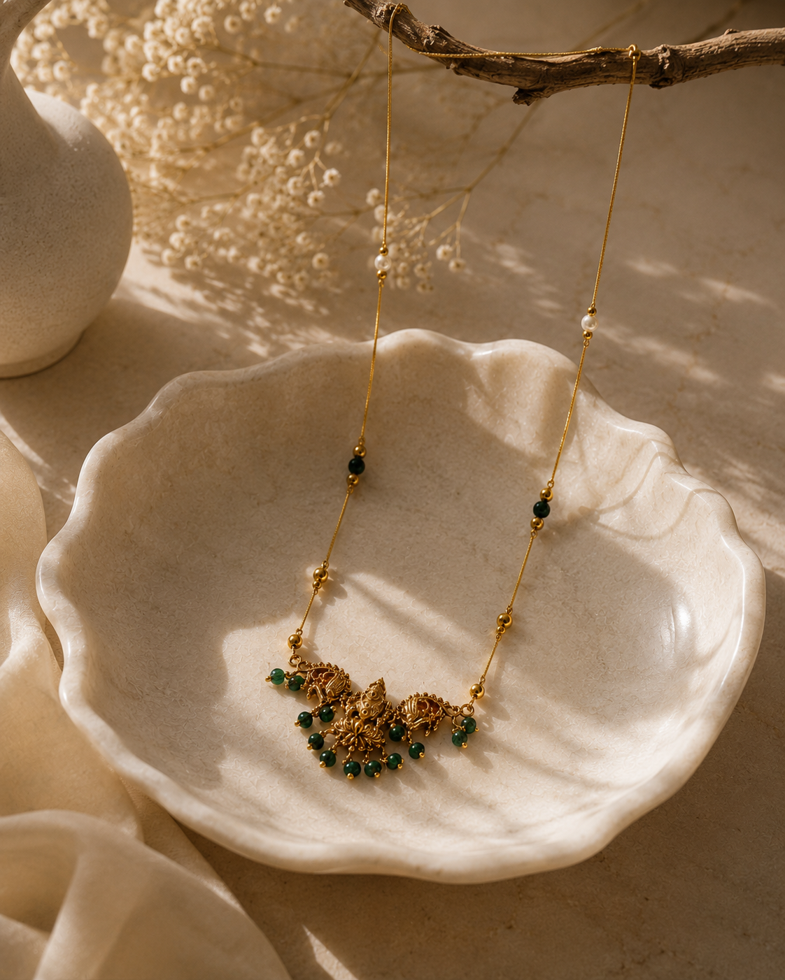 A gold necklace with a detailed floral pendant set with small green stones, displayed on a scalloped cream ceramic dish with warm natural light and dried flowers in the background.