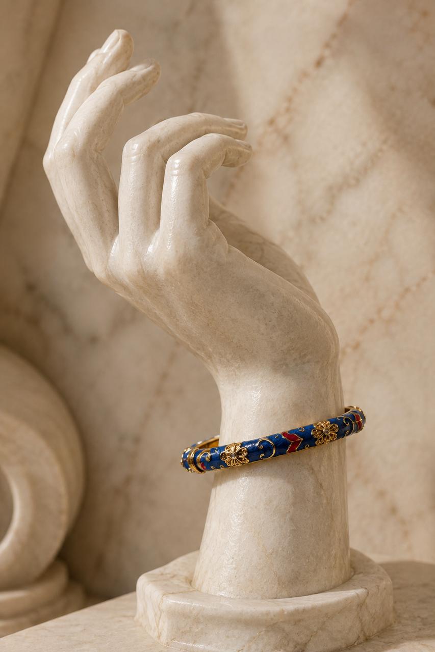 A gold and blue beaded bangle bracelet displayed on a cream-colored marble hand sculpture against a marble background.