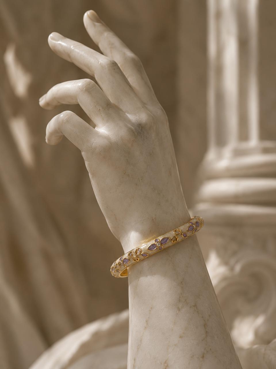 A gold bangle bracelet with purple and blue gemstones, shown on a marble hand sculpture in a warm, sepia-toned setting.