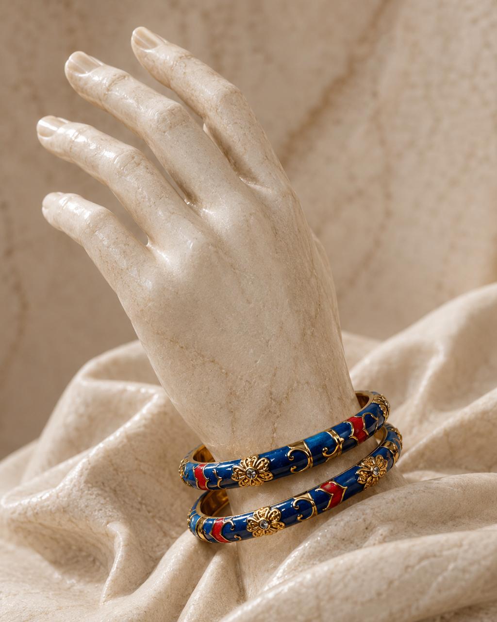 Two blue bangles with gold and red floral enameled details, displayed on a cream-colored stone hand mannequin over draped beige fabric.
