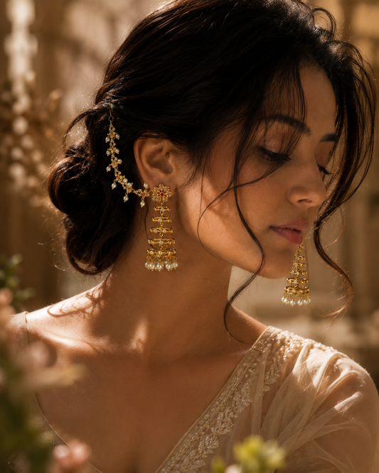 A woman with dark hair styled in an updo wearing ornate gold jhumka earrings with white bead accents, in warm golden lighting.