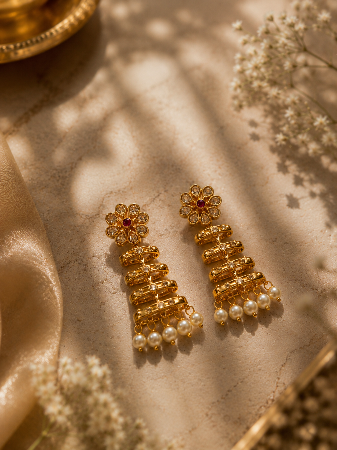 A pair of gold-toned traditional Indian jhumka earrings with floral tops, red stone centers, and dangling pearl beads, styled on a beige textured surface with soft shadows and dried flowers.