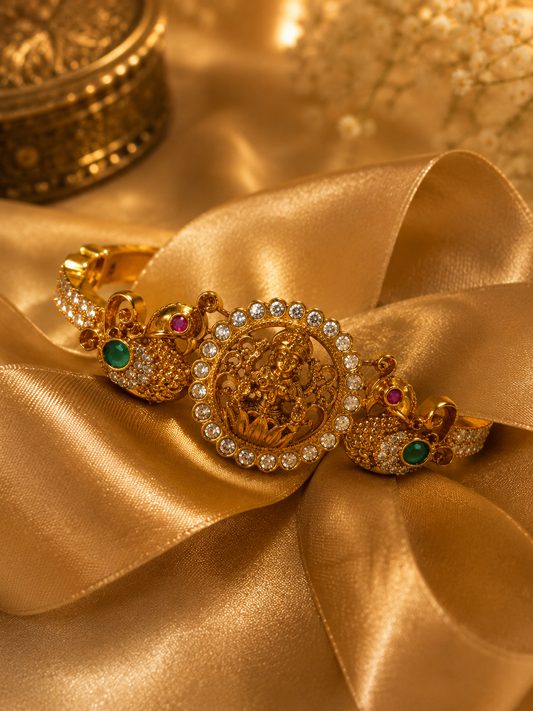 An ornate gold-toned jewelry piece displayed on draped satin fabric, featuring a central round medallion surrounded by small clear stones, flanked by two decorative side elements with green and pink stones.