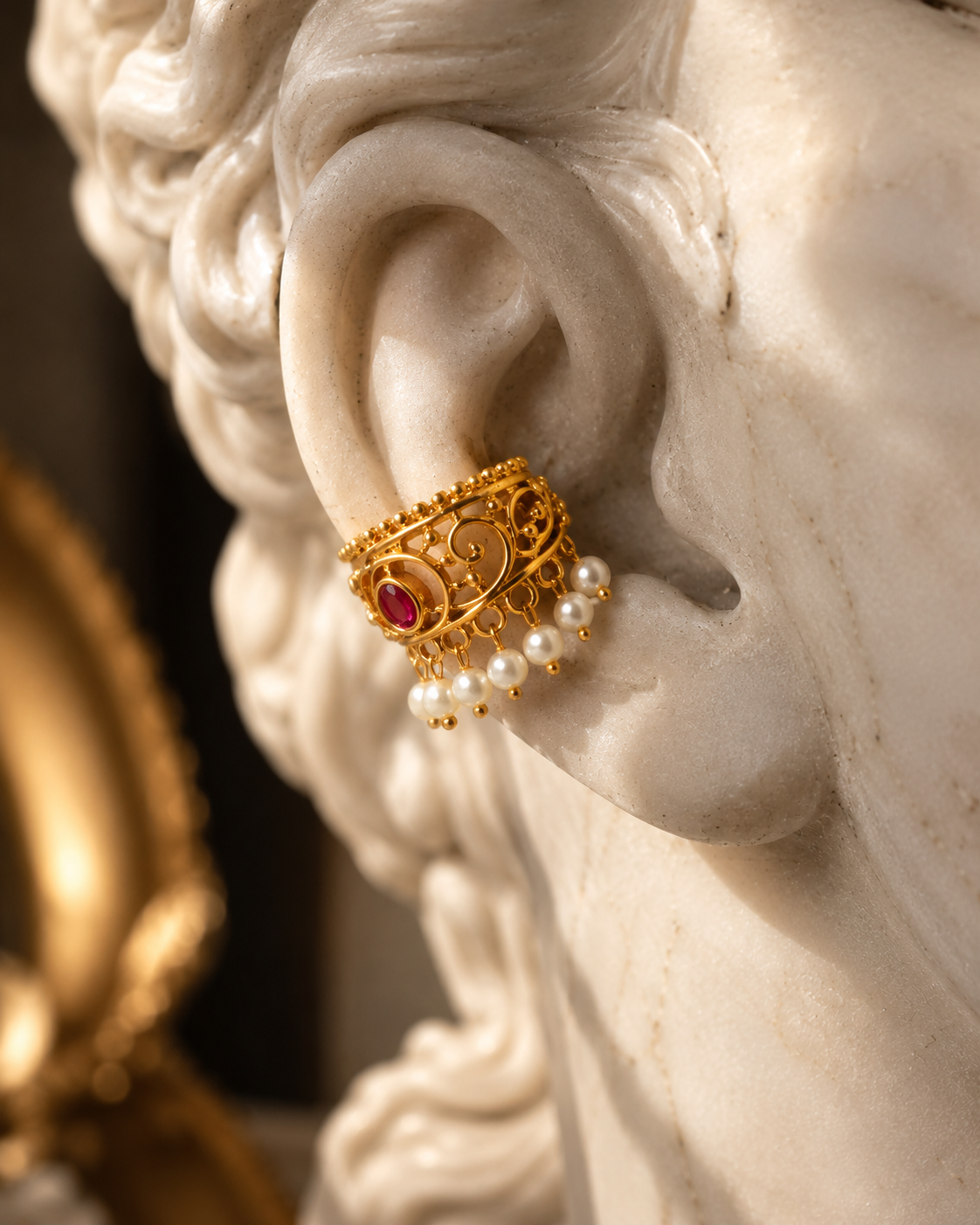 A close-up of a gold filigree ear cuff with a red gemstone and dangling white pearls, shown worn on a white marble statue's ear.