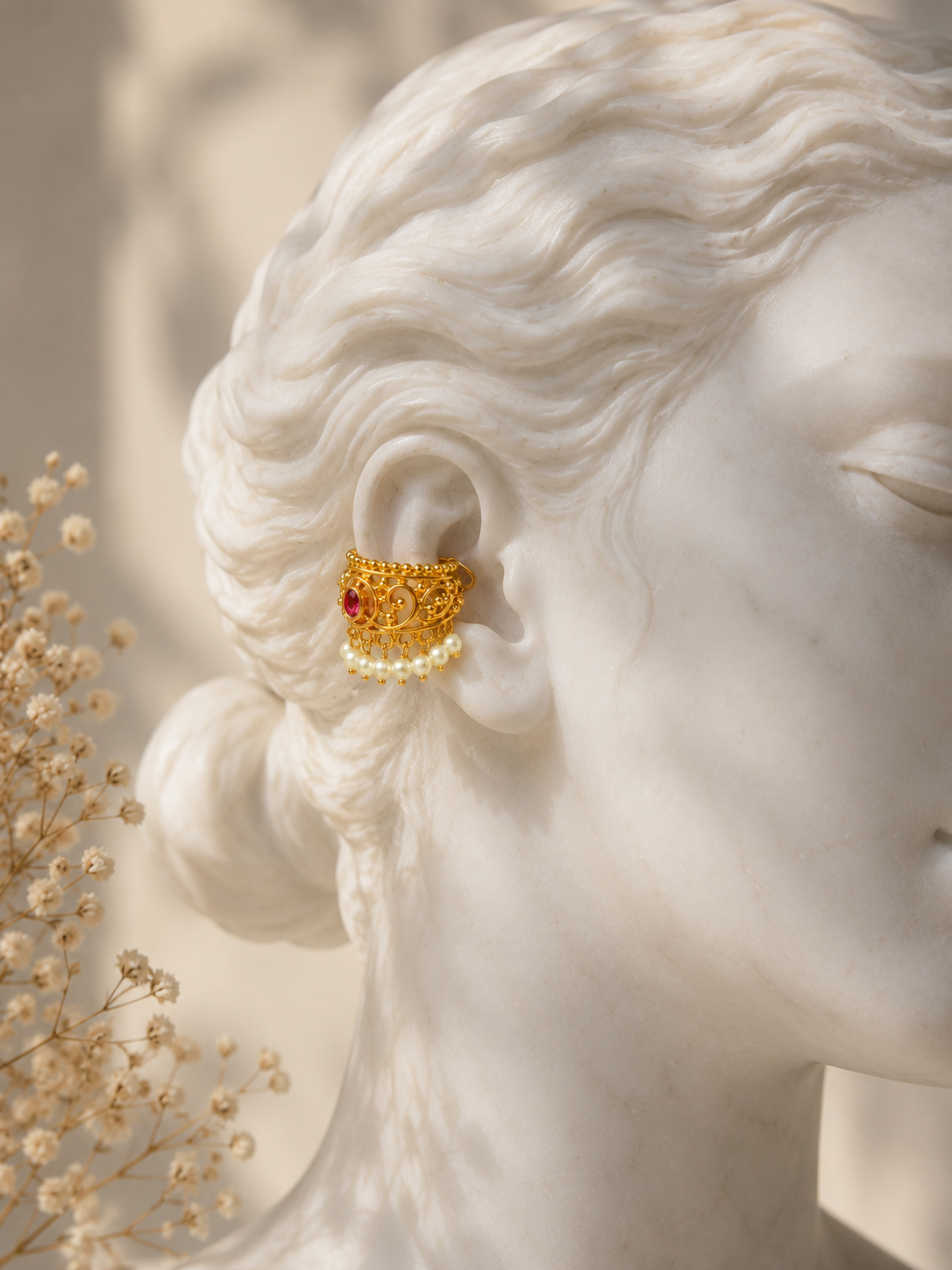 A gold ear cuff with a small red gemstone and a row of white pearls, shown worn on the ear of a white marble bust statue with dried flowers in the foreground.