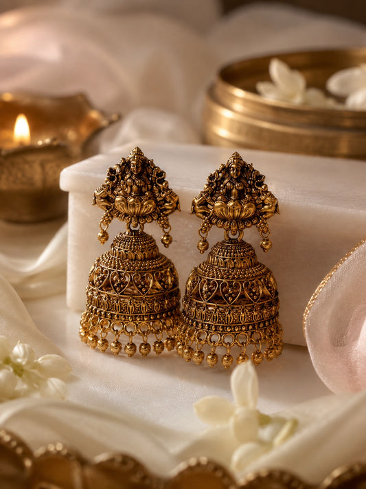 A pair of ornate gold-toned traditional Indian jhumka earrings displayed on a white jewelry box, surrounded by soft white fabric, small white flowers, and warm decorative lighting in the background.