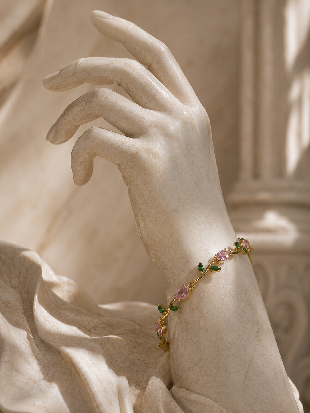 A delicate gold bracelet with pink and green gemstones is shown worn on a marble statue hand, set against a soft, classical stone background.