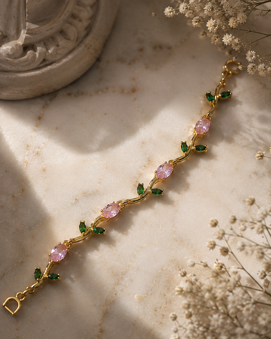 A gold-toned bracelet with oval pink gemstones and small green leaf accents, laid diagonally on a light marble surface with dried flowers and a decorative object in the background.